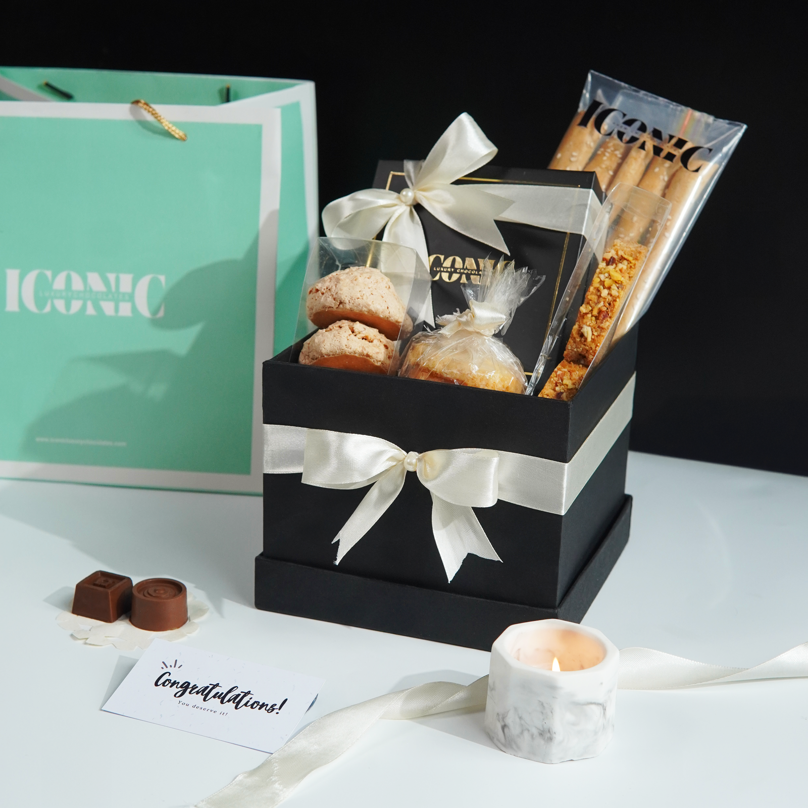 A black gift box filled with assorted cookies, chocolates, and breadsticks, tied with a white ribbon, sitting next to a turquoise shopping bag and a small candle.
