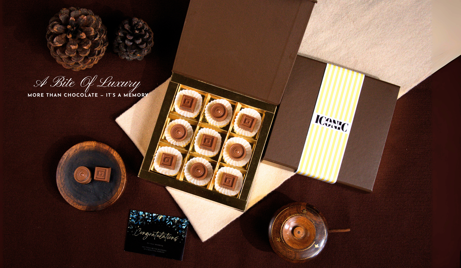 A brown box of nine chocolate squares with white bases, next to a "Congratulations" card, pine cones, and a small wooden dish.