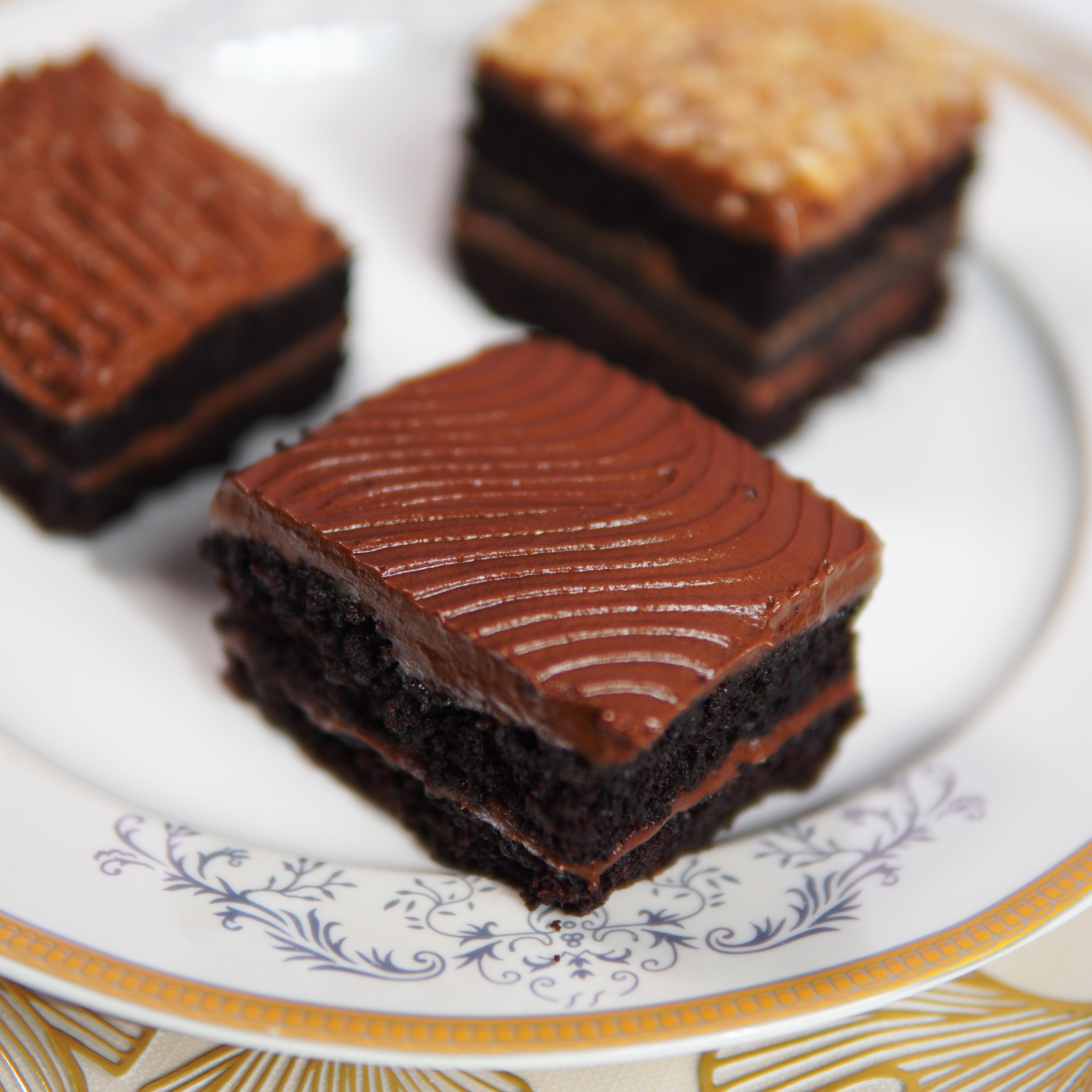 A plate featuring three square gourmet brownies. The closest brownie has a rich, dark chocolate frosting with decorative waves, while another in the background is topped with a textured caramel or praline layer.