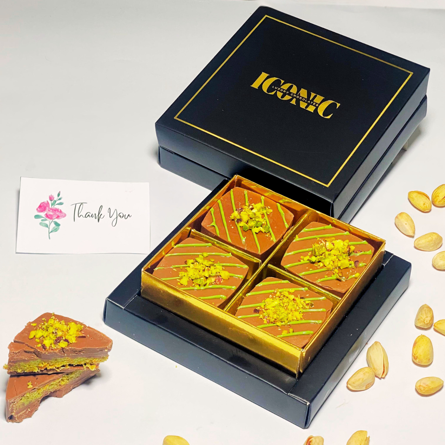 Elegant black and gold gift box containing four Pistachio Kunafa Mini Bars topped with crushed pistachios and green drizzle, with a thank-you card and pistachios scattered around.