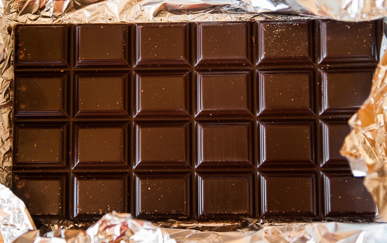 A bar of dark chocolate, partially unwrapped from its silver foil wrapper, showing a shiny, smooth texture.