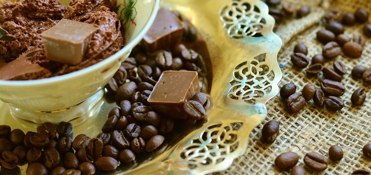 A creamy chocolate mousse dessert in a white bowl, with two small chocolate squares and scattered roasted coffee beans on a decorative gold tray and burlap surface.