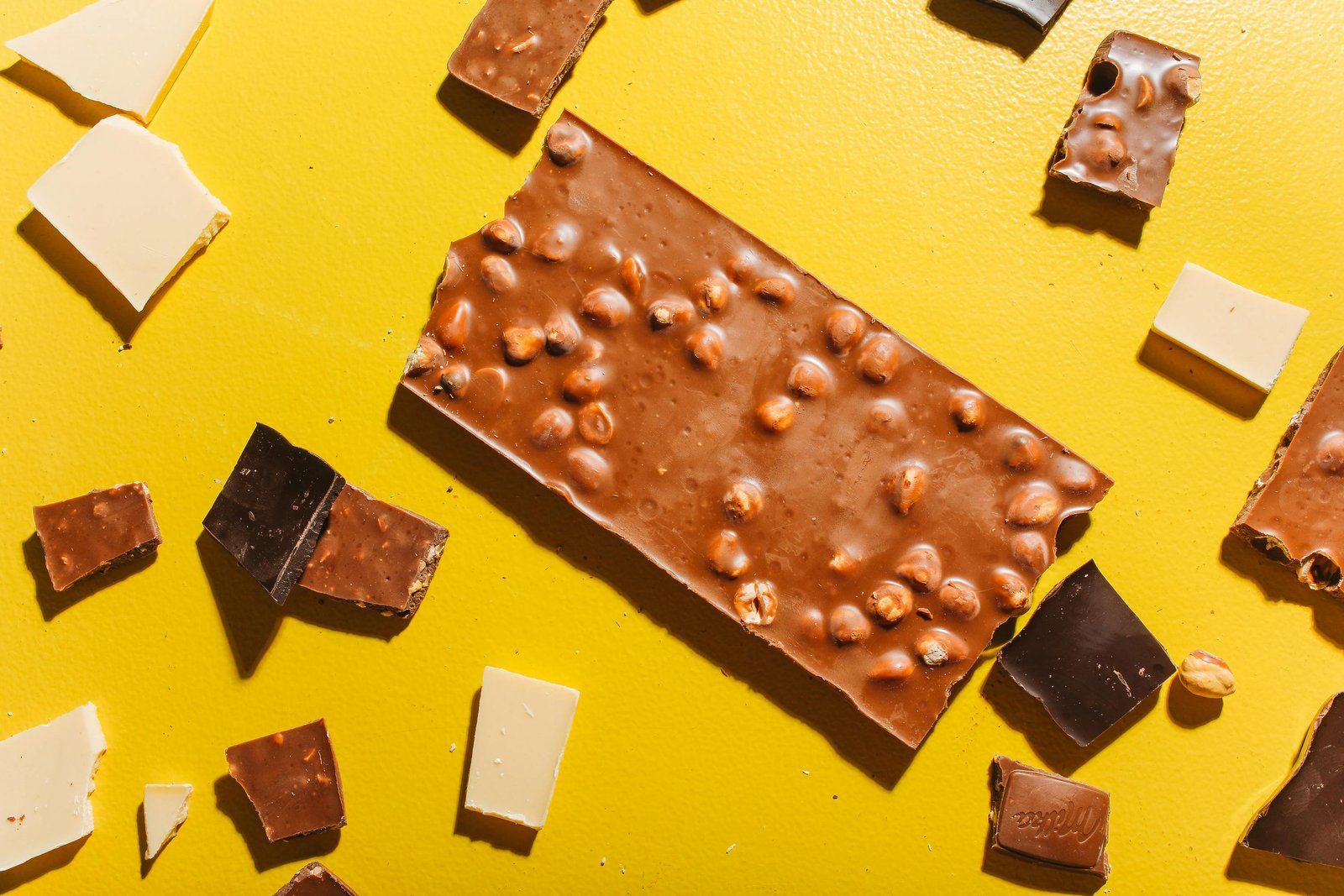 An image of various broken chocolate pieces on a vibrant yellow background. In the center is a large milk chocolate bar with whole hazelnuts. Around it are smaller chunks of milk, dark, and white chocolate.