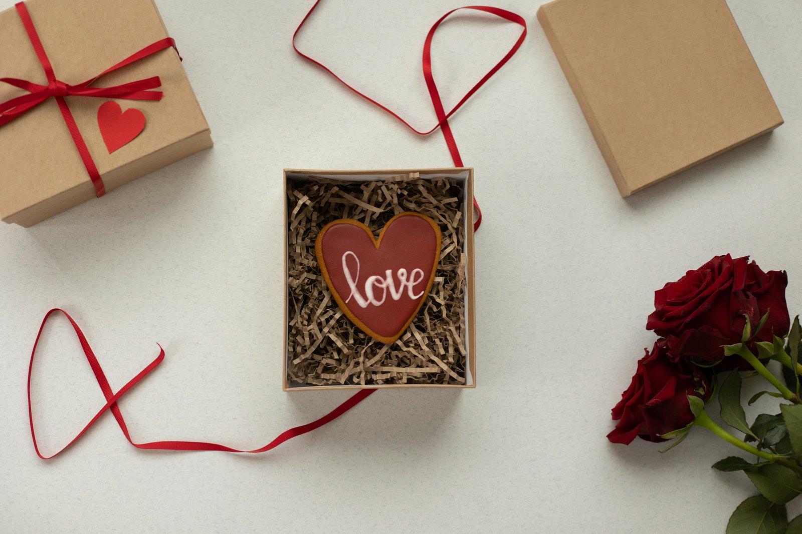 Heart-shaped chocolate cookie with the word “love” in a gift box with red ribbon and roses – symbolizing chocolate and romance.